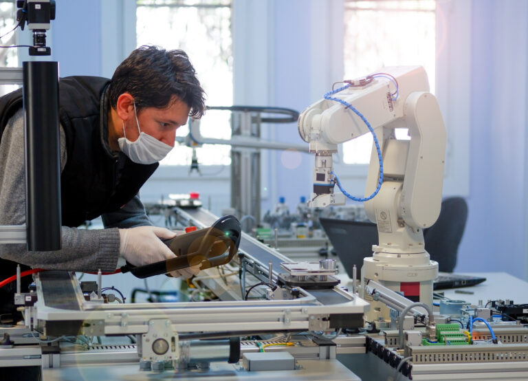 Man with surgical mask on face against COVID-19 ( Coronavirusis ) programming robot arm with control panel which is integrated on smart factory production line.