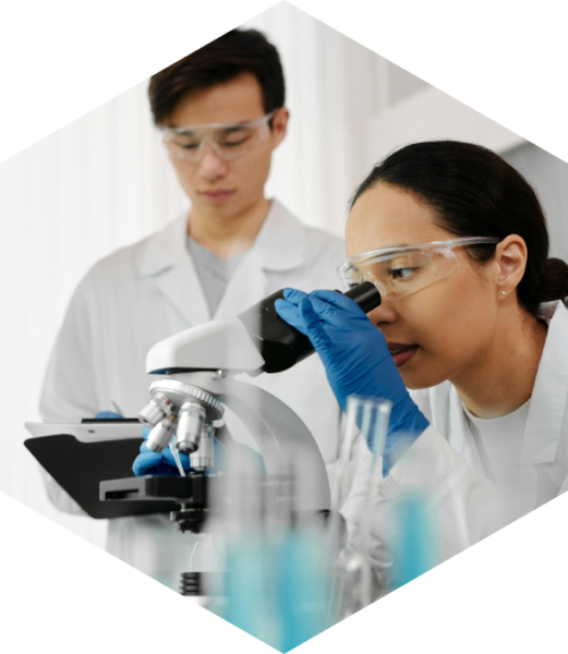Woman in a lab coat looking into a microscope while her lab mate is writing the data in the back